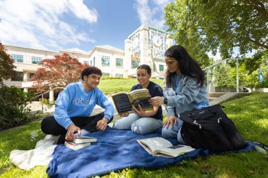 3 people sitting on a blanket, all looking at a book titled Season of the Witch.  The clocktower is behind them