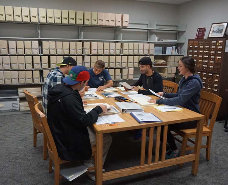 5 people sitting sound a table looking at documents.  There are walls of file cabinets behind them.