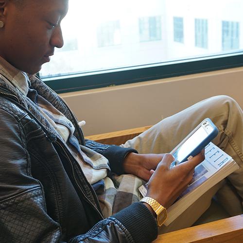 Young man sitting by a window holding a book and a smart phone and sending an email with his phone.