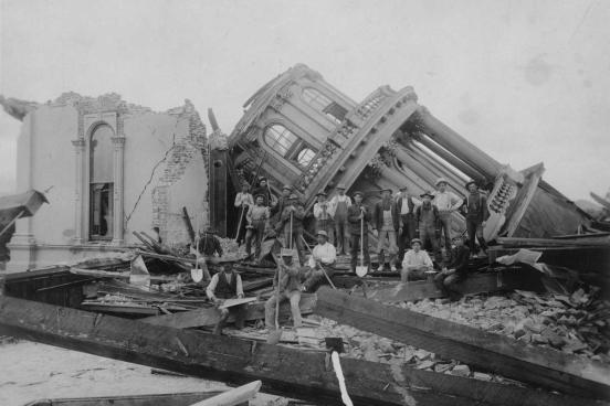 People standing in front of building destroyed by earthquake.