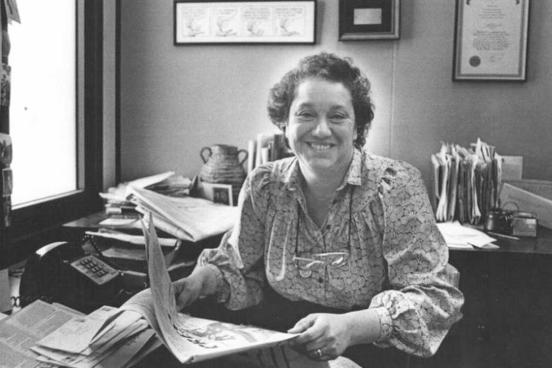 a woman holding a newspaper, sitting in an office. A typewriter and phone are behind her.