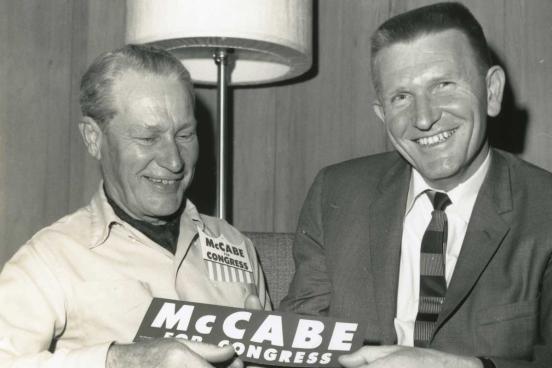 2 men, one wearing a McCabe for Congress button, both men hold a McCabe for Congress.bumpersticker