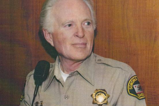 Man wearing a brown button up shirt with a badge and a sheriff patch on the right arm. 