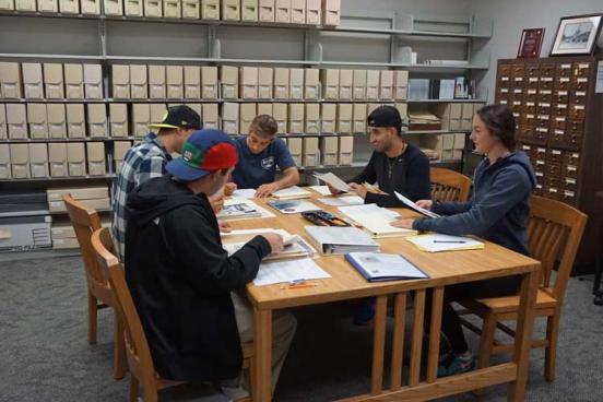 5 people sitting at a desk, looking at papers on the desk.