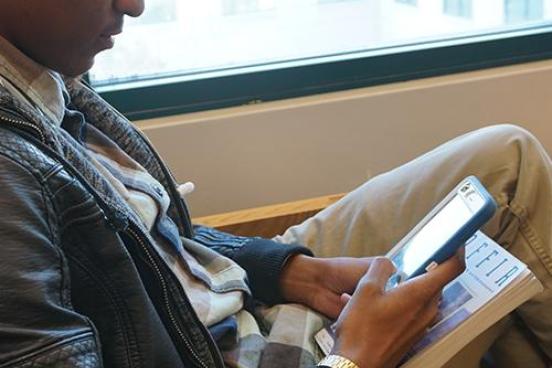 Young man sitting by a window holding a book and a smart phone and sending an email with his phone.