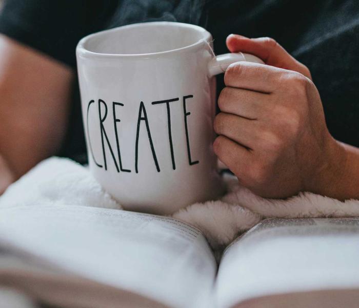 One hand holds the book, the other hand holds a mug that says create.