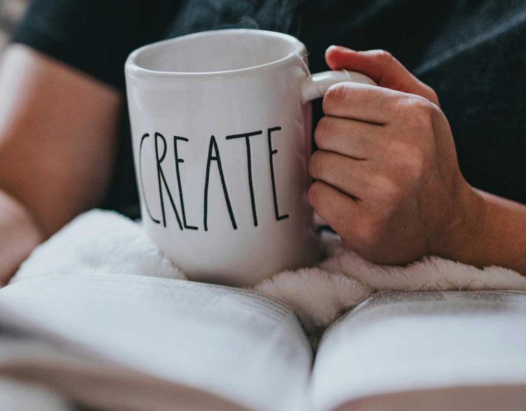 One hand holds the book, the other hand holds a mug that says create.