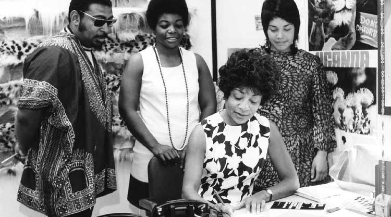 One woman is writing something 3 people stand behind her look at what she is writing.