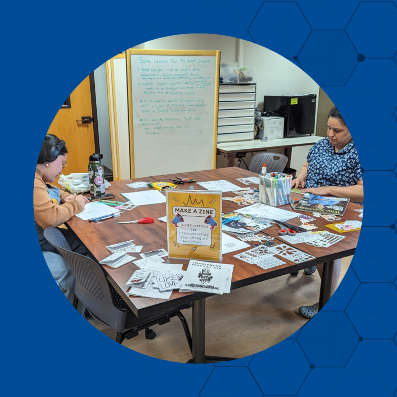 twp people sitting at a table.  A sign "Make a Zine" paper, pens and scissors are on the table.