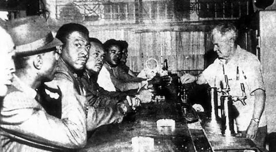 Six men sit at a counter on the left side.  On the right side, a man is serving them.