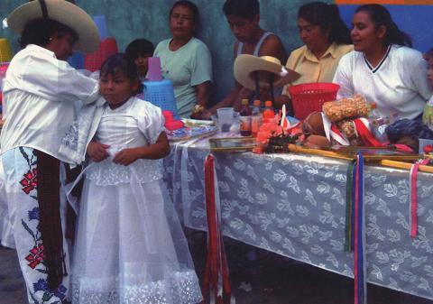 A woman wearing white pants with flowers braids a little girls hair while others watch.