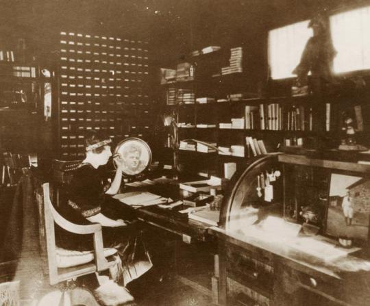 Woman sitting at a desk.  There are 2 pictures of Jack London on the desk.