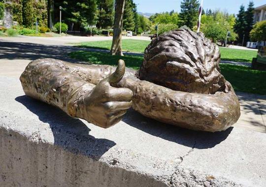Statue  of a persnn hiding their face and right hand is thumbs up.