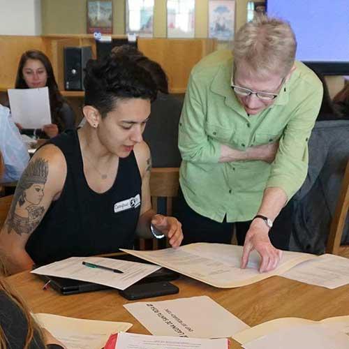 One woman sitting at a table, another woman is standing over her.  Both are looking at papers on the table.