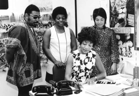 One woman is writing something 3 people stand behind her look at what she is writing.